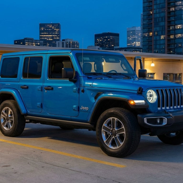 2025 Jeep Wrangler JL Unlimited in Hydro Blue Pearl at blue hour on a rooftop parking garage with city skyline, updated LED headlight signature, charcoal Hothead Headliner visible through the open passenger door