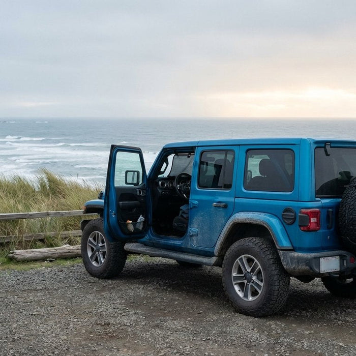 2020 Jeep Wrangler JL Unlimited in Ocean Blue Metallic at a coastal Oregon bluff rest-stop with driver door open showing charcoal Hothead Headliner, mesh trail bag loaded with a wetsuit and flip-flops on the spare, surfboard against the bumper